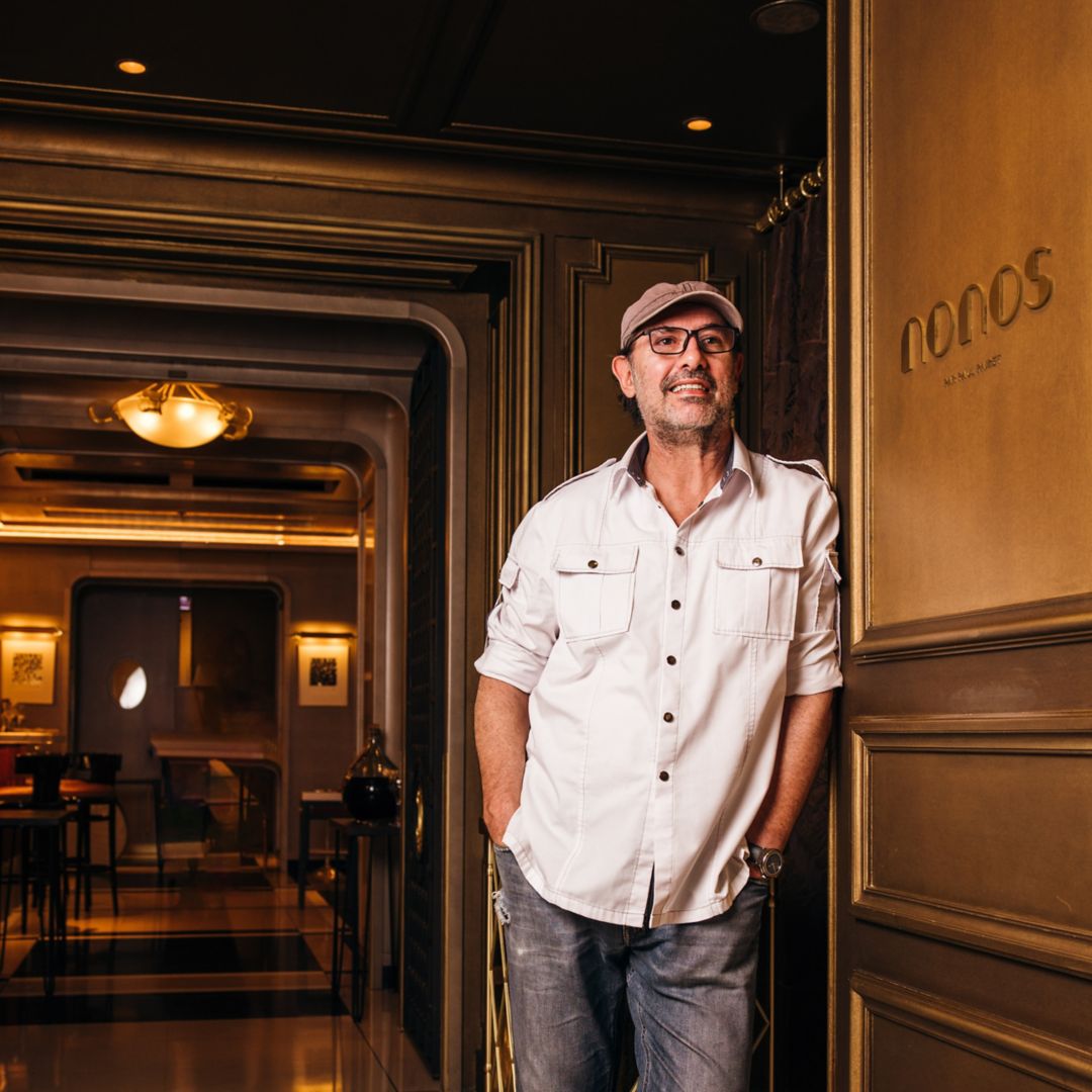 A man in a white shirt and cap standing in a doorway with a sign reading "Nonos" on the wall.